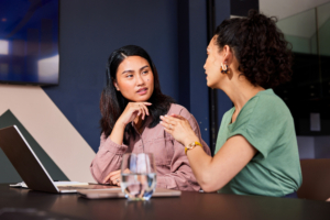 Adult businesswomen in their 30s sitting at desk with laptop, one woman explaining, co-worker listening with curious expression